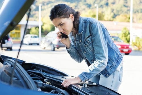 A woman inspecting her car’s engine with the hood open, calling for help on her phone after a breakdown.