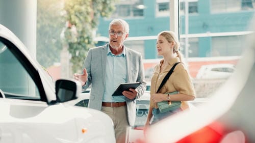 A salesperson showing a new car to a customer in a dealership showroom, explaining features while holding a tablet.