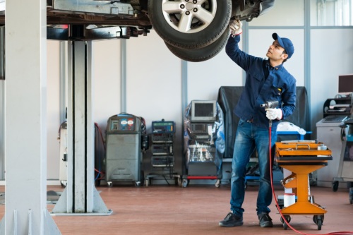 A mechanic working under a car on a hydraulic lift in an auto repair shop, using tools to inspect or service the vehicle.