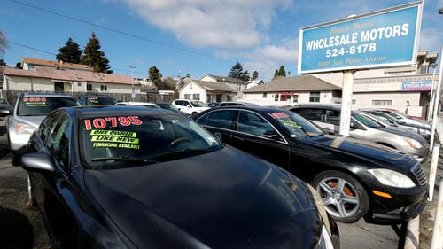 A used car dealership lot showing a black sedan with a price tag of $10,795 and signs like “LIKE NEW” and “FINANCING AVAILABLE.”