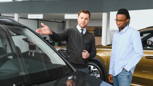 A car salesperson gestures toward a black car while explaining features to a customer in a dealership showroom.