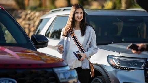 A woman points at a red Ford SUV while holding her phone and wallet, likely interacting with a car owner or staff member.