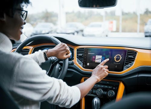 A driver interacting with a touchscreen infotainment system in a modern car with an orange-accented dashboard.