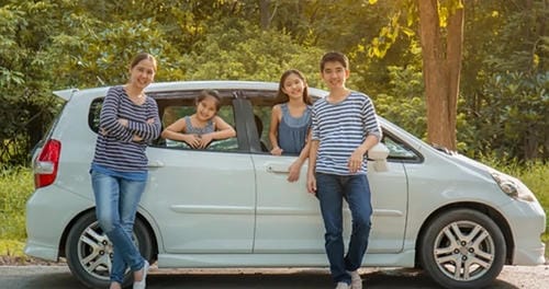 A family of four posing outside their white compact car in a wooded area.