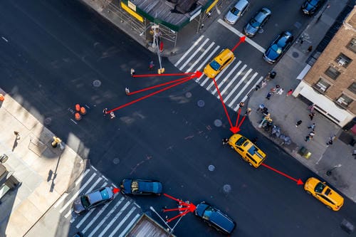 An aerial view of a city intersection with red lines tracking the movement of yellow taxis, illustrating traffic flow or autonomous vehicle path prediction.