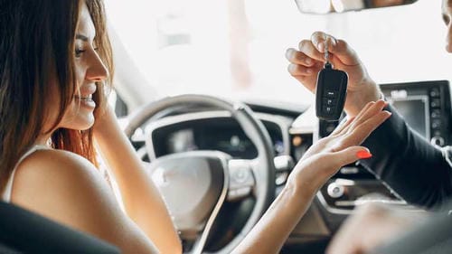 A woman receiving car keys from a salesperson inside a vehicle, marking the purchase completion.