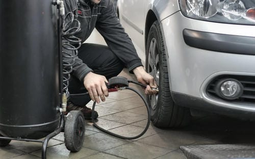 A mechanic using an air compressor and gauge to inflate or check the tire pressure on a silver car in a garage.