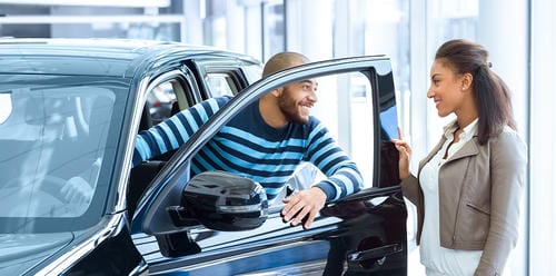 A man sitting in a new SUV, talking to a saleswoman at the dealership, likely preparing for a test drive.