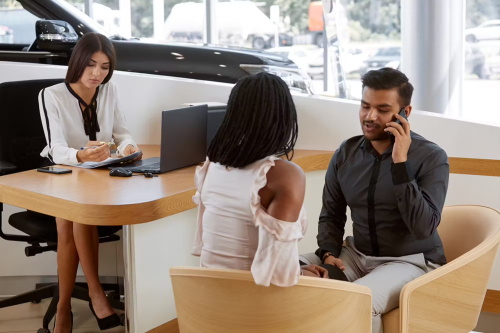 A salesperson, couple, and man discussing paperwork at a dealership desk, likely finalizing a car purchase or financing.
