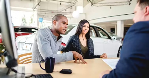 A couple sitting across from a sales representative at a dealership table, reviewing documents during the car-buying process.