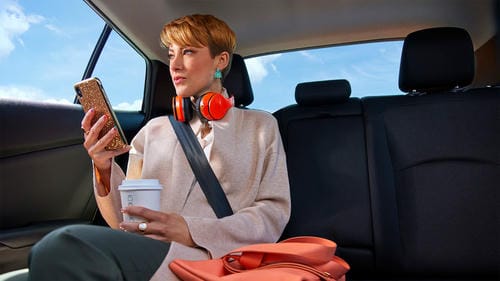 A passenger sitting in the backseat of a car, using her phone and holding a coffee cup.